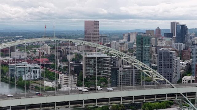 Steel arch bridge carries flowing traffic across Willamette River, framing downtown skyline rising beyond glass towers and historic buildings in Portland. Oregon, USA. Aerial drone view.