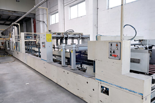 Industrial machinery for manufacturing corrugated cardboard boxes showing control panel and conveying section inside a production plant.