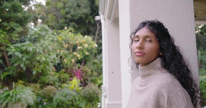 African American woman leaning on porch column in beige turtleneck, gazing at trees, smiling