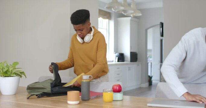 Morning African American dad and teen son packing backpack, sliding laptop into bag at kitchen bar