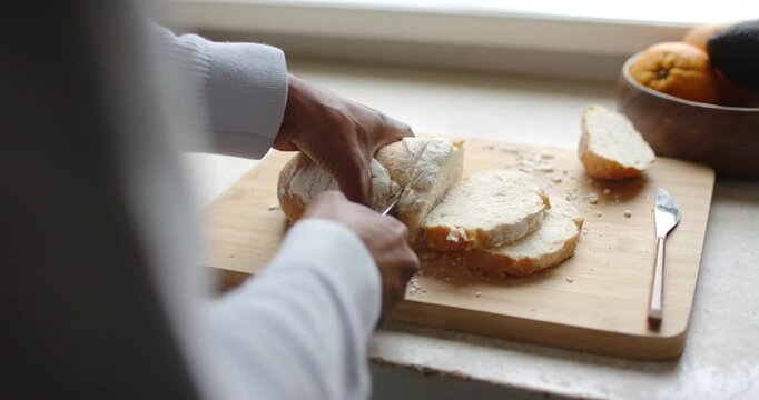 African American man holding loaf, slicing bread with bread knife on board by window preparing food