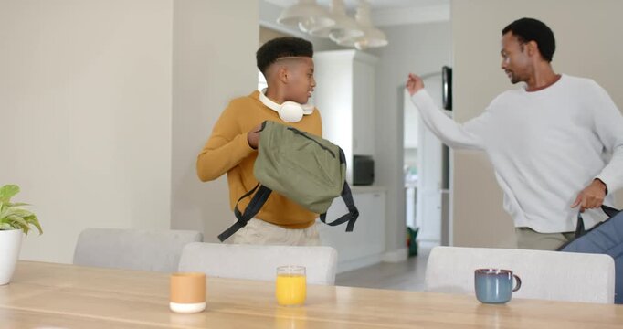 African American dad after gesture guiding teen son adjusting green backpack at table, toward hall