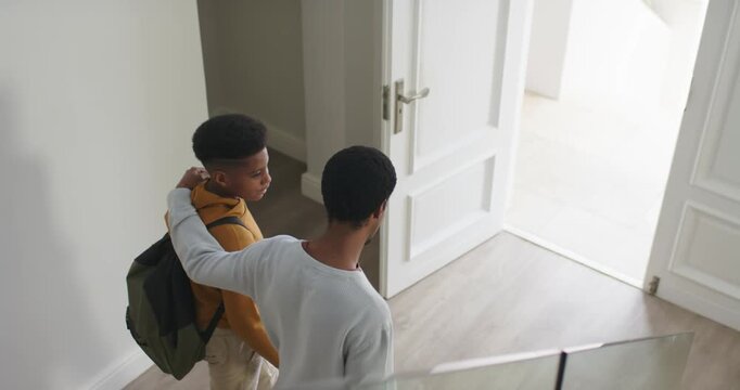 African American dad arriving behind teen in foyer, steadying backpack guiding him toward open door