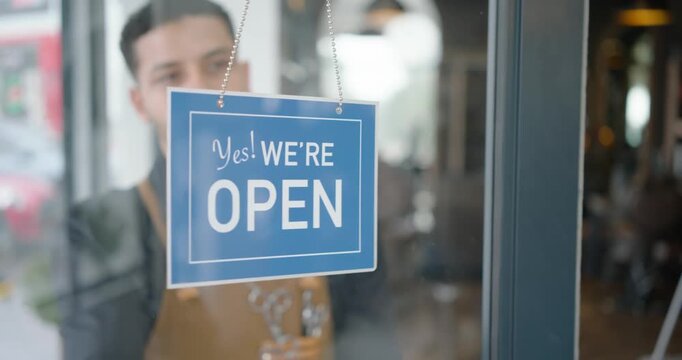 Owner is reaching, unhooking red CLOSED sign on door and hanging blue OPEN sign welcoming customers