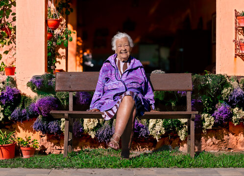Smiling elderly woman sitting on a wooden bench in a sunny garden surrounded by purple flowers, enjoying retirement and wellbeing.