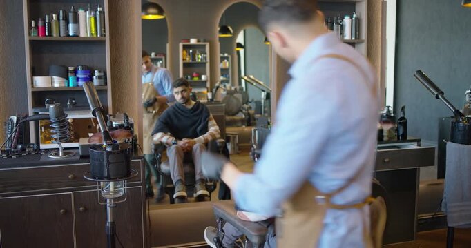 Aproned male barber trimming and drying two seated men's hair with clippers, finishing cut in shop