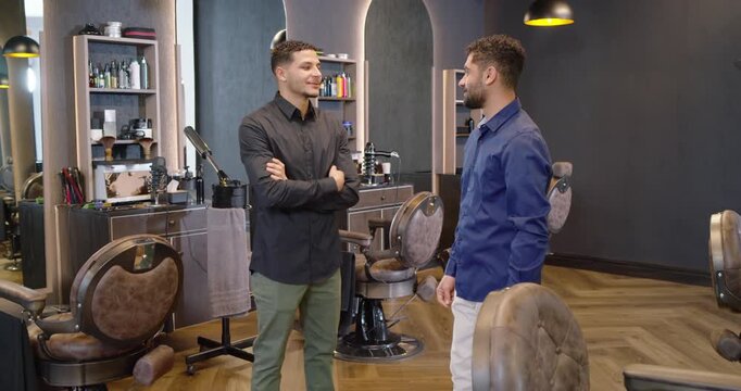 Two male partners in smart shirts conversing at barbershop near chairs shifting posing for portrait