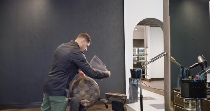Male barber entering salon corner wiping chair with cloth readying station for client, copy space