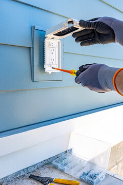 Side view of electrician hands using a screwdriver to install a weatherproof outdoor outlet cover on a blue siding house wall