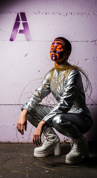 A person in a silver outfit with face paint squats against a textured wall with a stenciled letter A, representing futuristic fashion or artistic expression
