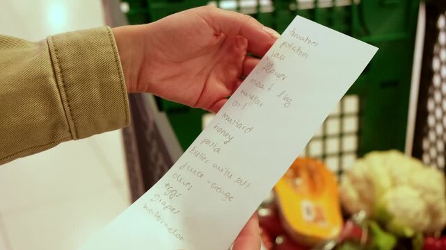 Unrecognizable woman checking a handwritten shopping list while standing with a trolley full of fresh vegetables in a grocery store, making sure she has everything she needs for her recipe