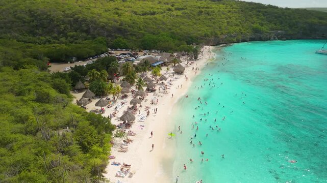 Aerial orbit above busy Playa Cas Abao revealing white sand, thatched palapas, swimming tourists and catamaran in turquoise Caribbean bay of Cura&ccedil;ao