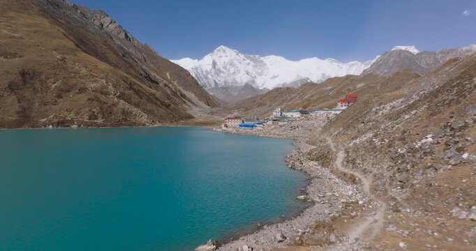 Gokyo Lake, Everest Region, Nepal drone push-in shot over the turquoise water toward the lakeside settlement and the snow-covered peak of Cho Oyu mountain