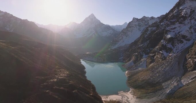 Chola Tshe Lake Everest region Nepal, aerial drone shot showing the snow-covered peak of Ama Dablam mountain rising behind hills with the sun beaming into the frame over the water surface