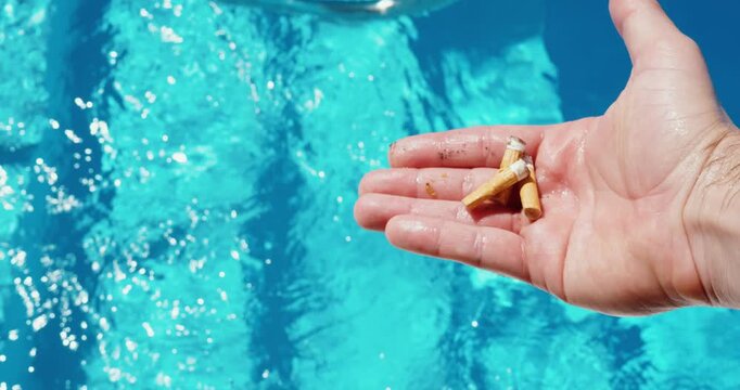 Close-up of a hand holding cigarette butts next to a swimming pool, emphasizing strict no-smoking rules and environmental protection around pool areas.