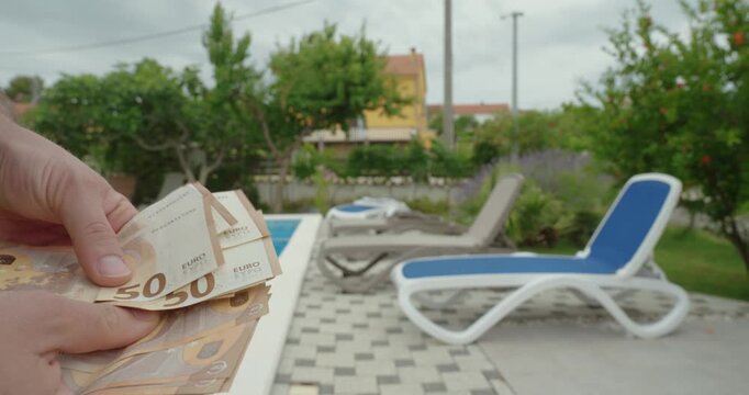 Close-up of hands counting euro banknotes near a swimming pool and sun loungers, representing vacation budgeting, travel expenses, holiday planning, personal finance, savings, and leisure spending dec