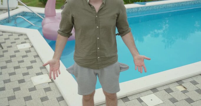 Young man standing beside a swimming pool with turned-out empty pockets, symbolizing lack of money, financial difficulties, budgeting, expenses, debt, economic challenges, and personal finance concept