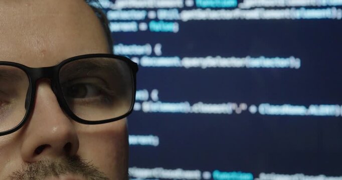 Close-up of a man wearing glasses while looking at a computer screen with programming code in the background. Technology, software development, concentration, and digital work concept.