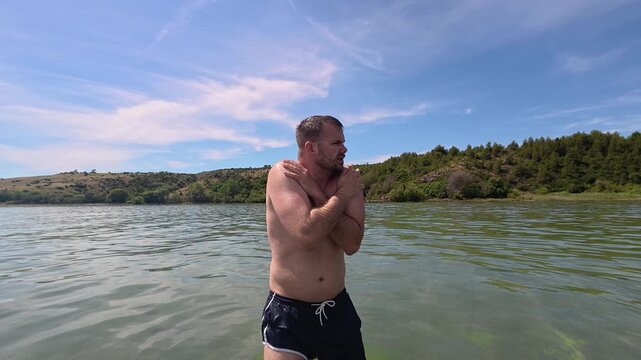 Man standing in shallow sea water with crossed arms, reacting to cold temperatures. Outdoor summer scene illustrating chilly water conditions, discomfort, and natural coastal recreation.