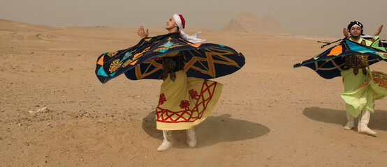 Fototapeta premium Two Egyptian Tanoura folk dancers spinning in vibrant flared costumes before the Giza pyramids in the desert ideal for cultural festival tourism and