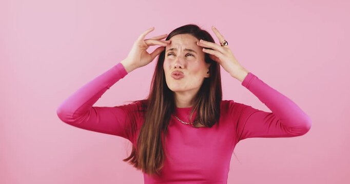 Young woman feeling stressed and overwhelmed, suffering from headache and intense frustration, expressive facial gesture of suffering and emotional distress studio portrait isolated on pink background