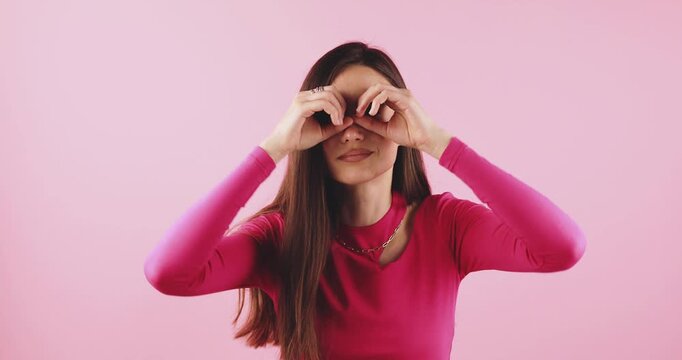 Young woman looking around and making binoculars gesture with hands over eyes, expressive and playful curiosity, studio portrait isolated on pink background, concept for searching, observing