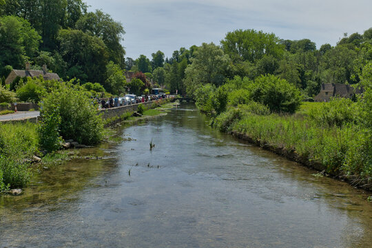 A picturesque view of the River Coln flowing through Bibury, England, featuring clear water, ducks, green trees, and traditional stone buildings.