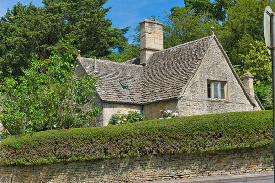 A charming stone cottage with a grey slate roof and tall chimney, set behind a neat hedge. This picturesque English scene is surrounded by green trees in Bibury - Gloucestershire - UK