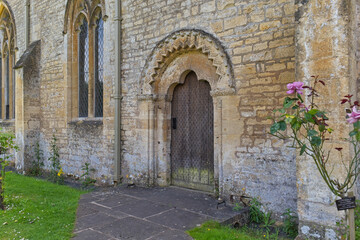 Naklejka premium A historic stone building features an arched doorway with a weathered wooden door. Green grass and a pink rose bush flank the entrance to the old structure in Bibury - Gloucestershire - UK