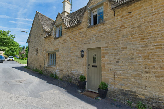 A quaint, light-colored stone cottage with a grey door and potted plants lines a sunny village street in Bibury - Gloucestershire - UK