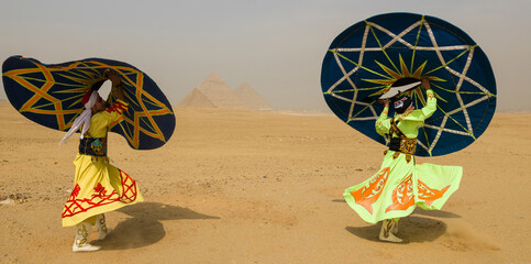 Fototapeta premium Two vibrant Tanoura folk dancers spinning colorful star patterned skirts overhead in the desert before the Giza pyramids in Egypt ideal for tourism