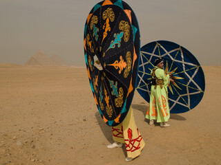 Fototapeta premium Vibrant Egyptian tanoura folk dancers carrying their colorful spinning Sufi skirts across the desert sand with the Giza pyramids behind ideal for