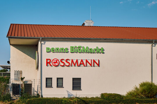 Denns BioMarkt and Rossmann logos on a white building wall with a brown roof in Fuerth, Germany, under a clear sky.