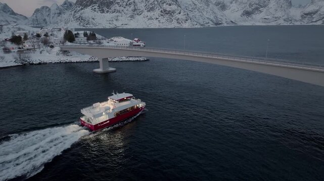 Red and white passenger ferry passing under curved bridge near snowy coastline at dusk