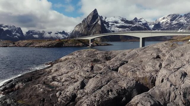Rocky coastal fjord with bridge, lone hiker, and snowy mountains under cloudy sky