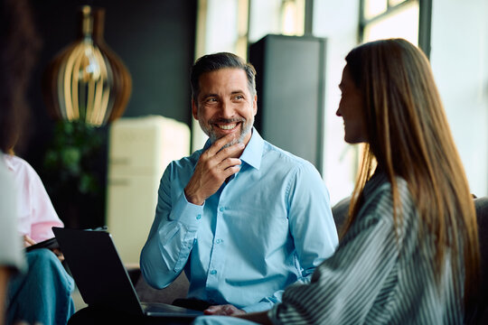 Happy businessman communicating with colleague while working on laptop in office.