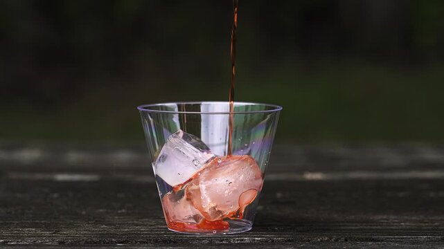 Refreshing red beverage pouring into a clear plastic cup filled with ice cubes. The cup sits on a dark wooden picnic table outdoors in slow motion