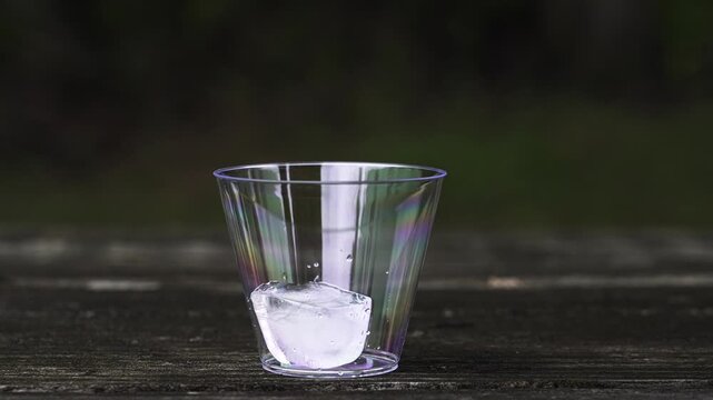 Slow motion close-up view of a person's hand placing ice cubes into a clear plastic cup resting on an outdoor wooden table for a refreshing drink