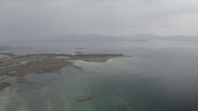 A stunning aerial view of Lake Sevan in Armenia, featuring a winding sandy peninsula, small lakeside settlements, and marshy wetlands under an overcast, cloudy, and atmospheric autumn sky.

