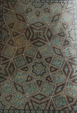 Close-up of a traditional Persian geometric inlay panel serving as a textured background, featuring an intricate interlocking star pattern with finely dotted borders in faded turquoise and brown tones