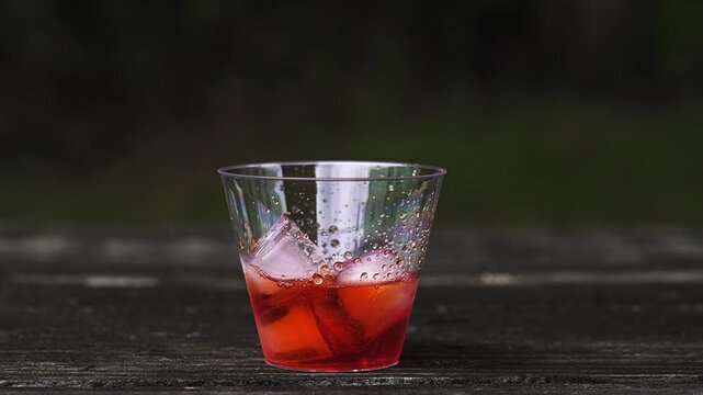 Refreshing spritz cocktail being prepared on a rustic wooden table. Bartender adds soda and garnishes the red drink with a fresh orange slice in slow motion