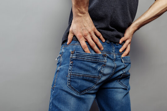A young, thin guy in jeans is squeezing his ass tightly with his hands. Concept: Queue to the bathroom, intestinal and stomach diseases. expressive veins on the arms