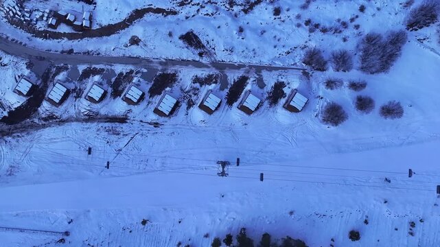 Aerial views of a winter ski resort with a chairlift operating on snowy mountain slopes against a beautiful pink sunset sky, offering a scenic and serene alpine landscape.

