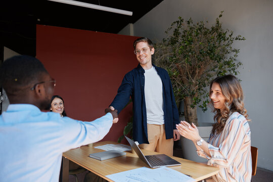Two male business people of different ethnicities shaking hands. Forming a partnership deal. While a female colleague claps enthusiastically