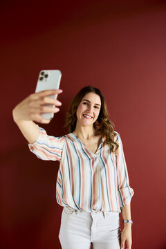 Young woman smiling while capturing a selfie using a mobile phone, demonstrating self-portrait photography and content creation on a red background