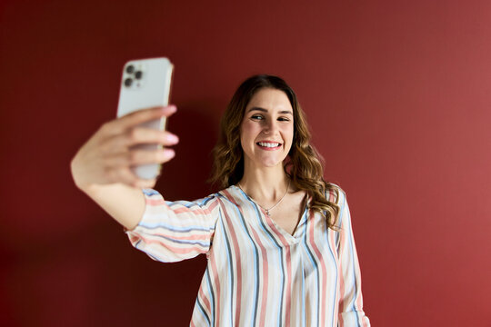 Young smiling woman holding modern smartphone taking selfie, filming video blog, or video calling on social media against a vibrant red background