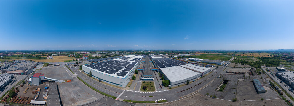 Aerial view of a large industrial factory complex featuring extensive solar panel arrays on the rooftops, surrounded by paved roads and open fields in Chivasso, Piedmont, Italy.
