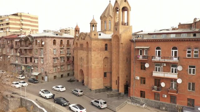 An aerial drone shot reveals Saint Sarkis Cathedral in Yerevan, Armenia, standing on the edge of the Hrazdan Gorge, surrounded by historic tufa buildings on a cloudy day.

