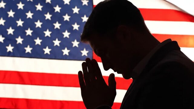 Silhouette of businessman in suit praying with hands together in front of american flag.