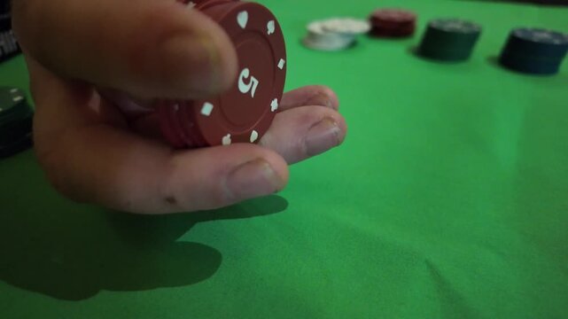 Close up view of an individual's hand idly handling red poker chips against a green table background. The mood is relaxed, yet engaged, suggesting a casual game or perhaps anticipation. The setting is